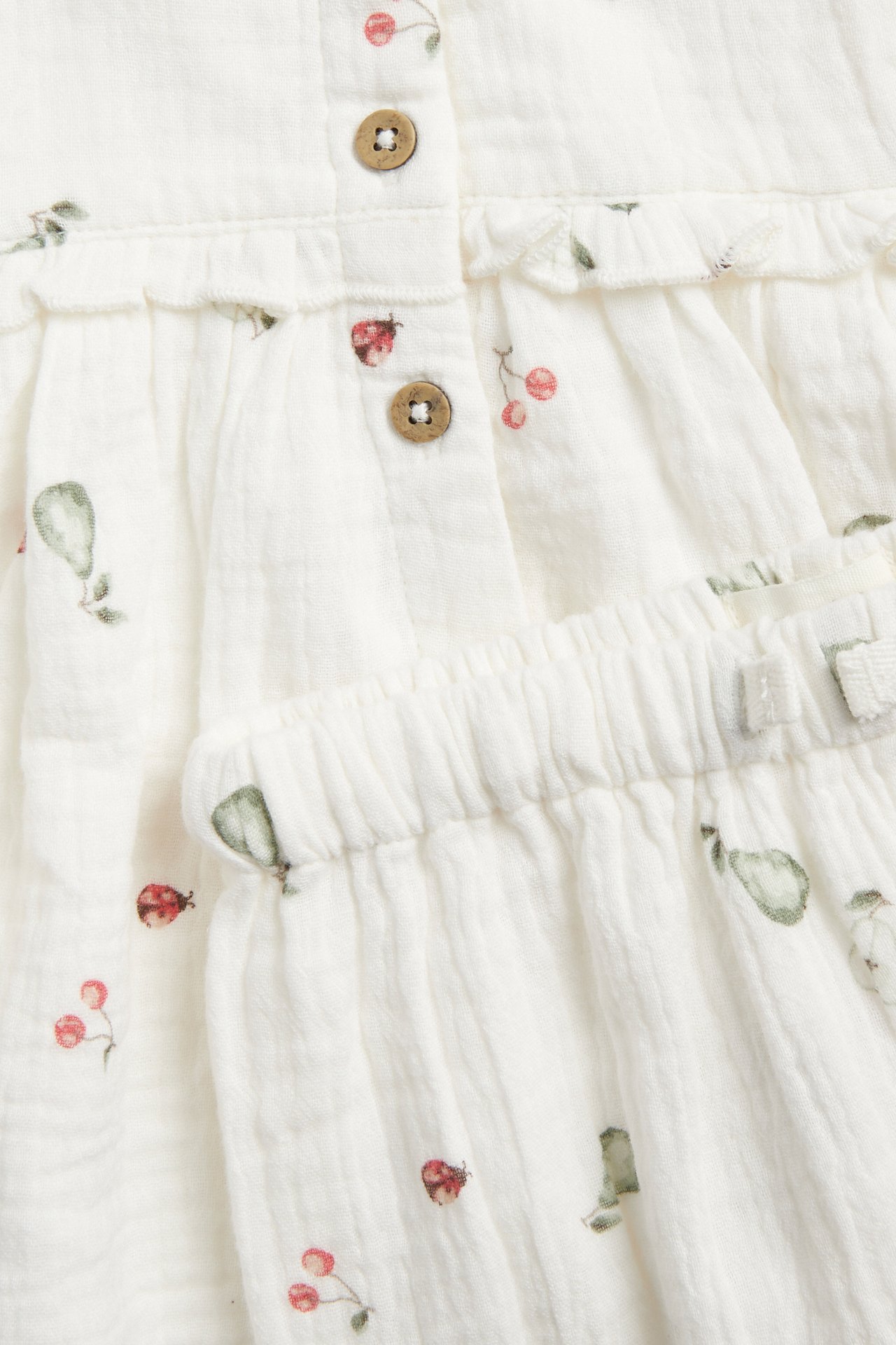 Close-up of an off-white muslin set with pear, cherry, ladybug print, buttons, and ruffles.