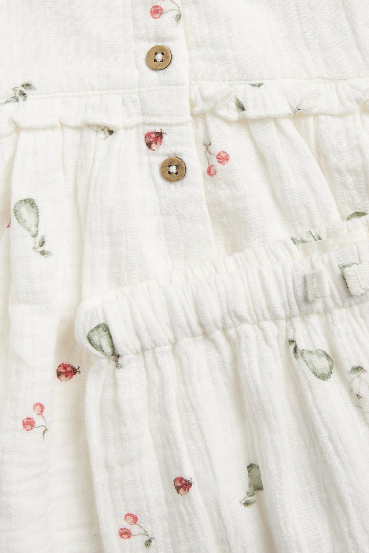 Nahaufnahme eines cremeweißen Musselin-Sets mit Birnen-, Kirsch- und Marienkäfer-Muster, Knöpfen und Rüschen.