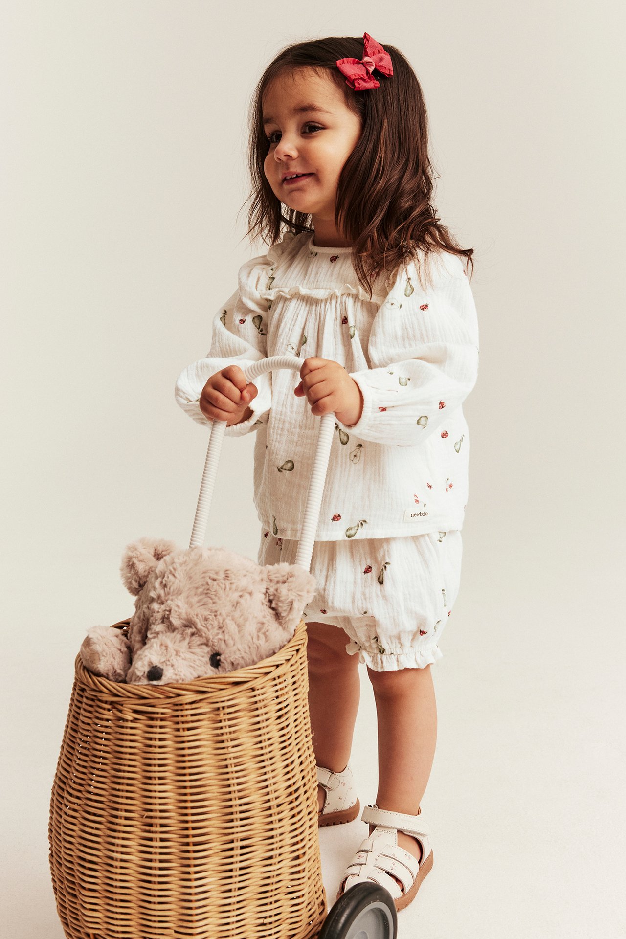 Front view: Kid in off-white patterned muslin set, red bow, white sandals, holding a basket.