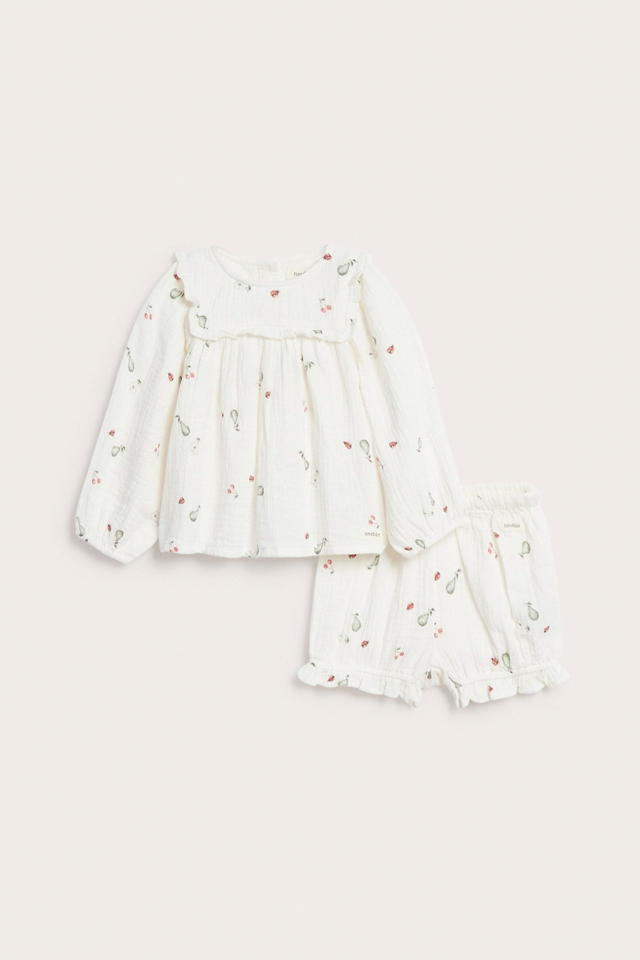 Vorderansicht eines cremeweißen Musselin-Baby-Sets mit langärmeliger Bluse mit Rüschenpasse und gemusterten Shorts.