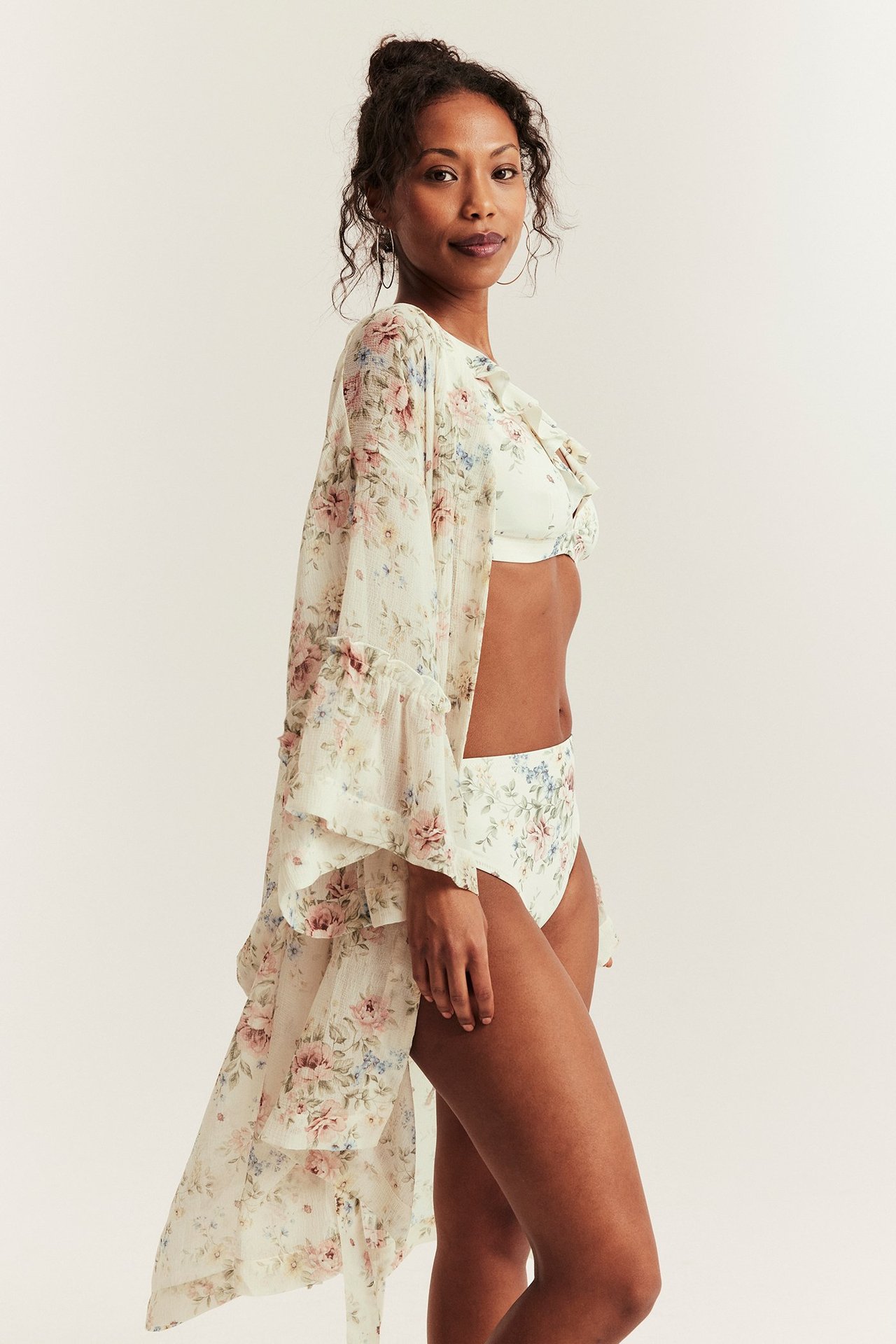 Seitenansicht einer Frau in cremeweißen floralen High-Waist-Bikinihosen, passendem Bikinitop und langem, fließendem Kimono.