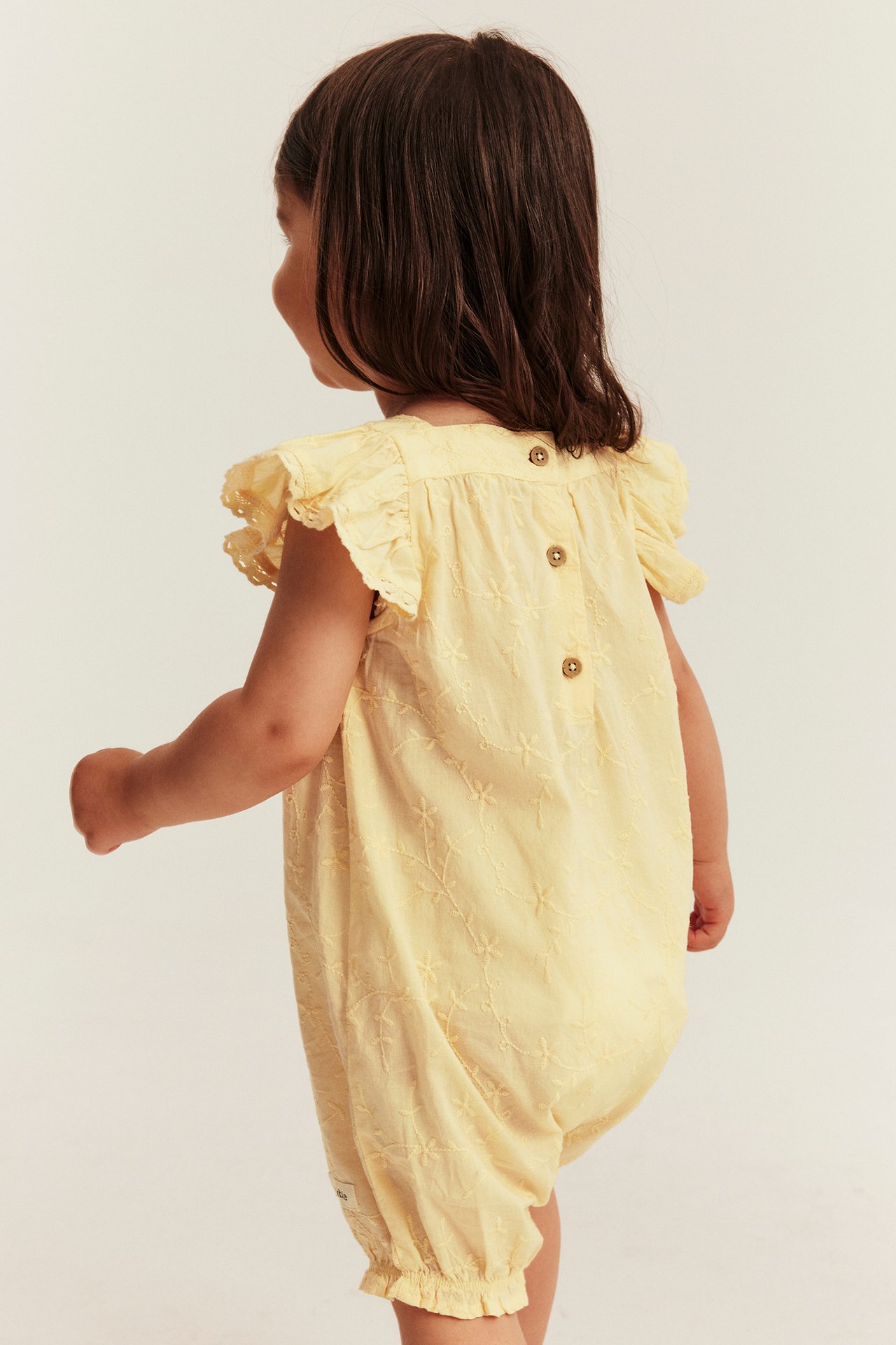 Back view of a baby in a yellow embroidered cotton romper with ruffled lace sleeves and back buttons.