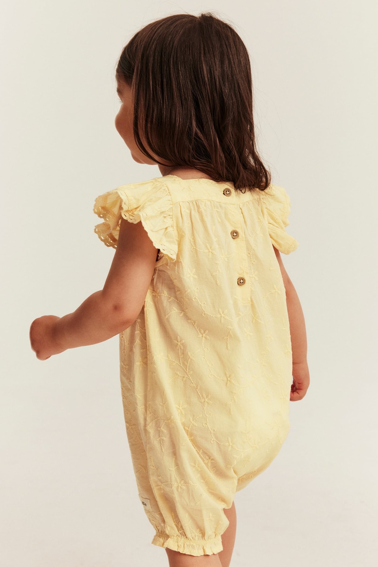 Rückansicht eines Babys in gelbem bestickten Baumwoll-Romper mit Rüschenärmeln und Knöpfen am Rücken.