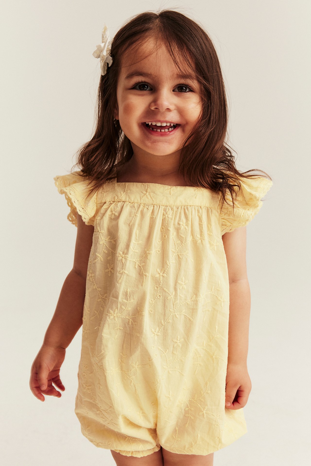 Front view of a smiling kid wearing a yellow embroidered woven short-legged romper with ruffle sleeves and a white hair clip.