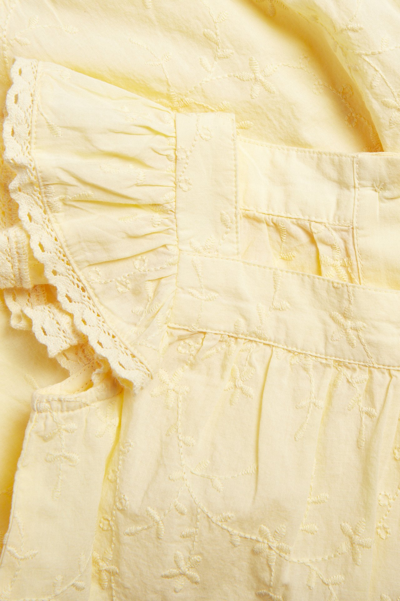 Close-up front view of a baby's yellow woven cotton romper with embroidered pattern and ruffled sleeves with lace trim.