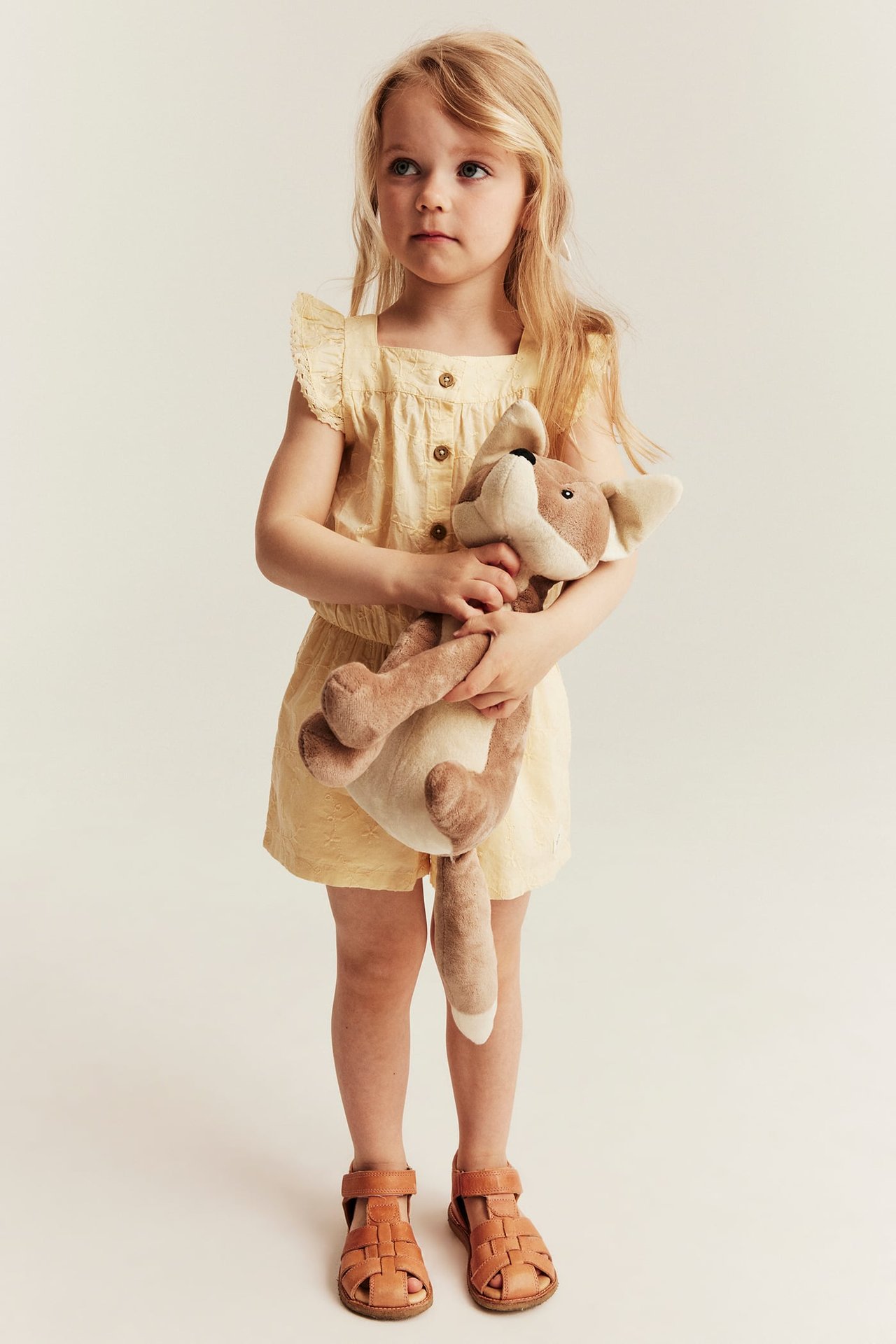 Vorderansicht: Kind trägt einen hellgelben bestickten Kurz-Jumpsuit mit Rüschenärmeln und braunen Sandalen, hält einen Stoffhund.