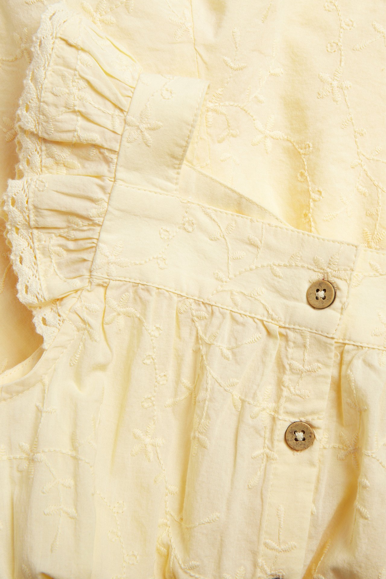 Close-up of a light yellow embroidered woven jumpsuit with ruffle sleeves, lace trim, and front buttons.