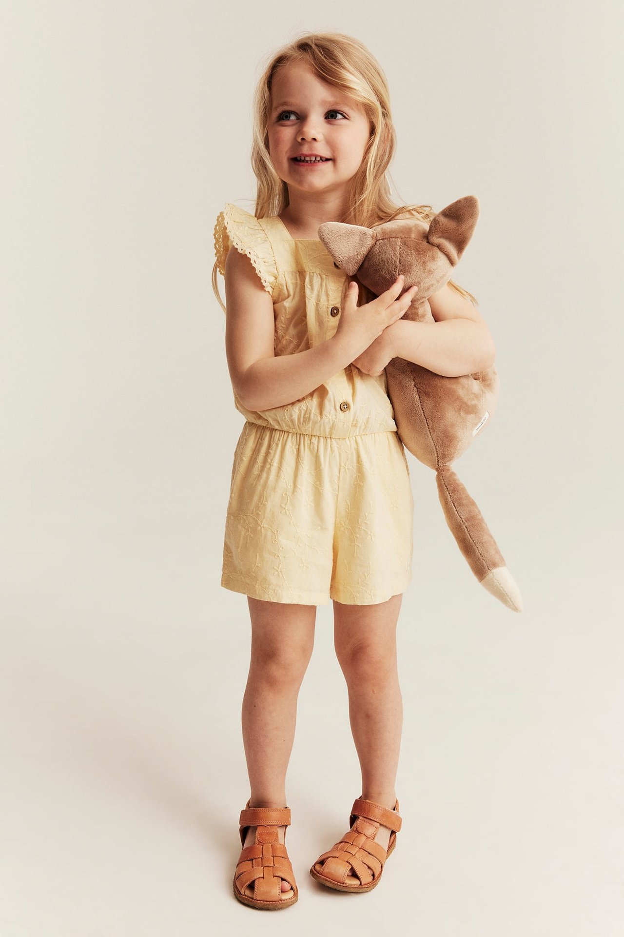 Vorderansicht: Kind in gelbem besticktem Kurz-Jumpsuit, Rüschenärmeln, hält braunes Stofftier, braune Sandalen.