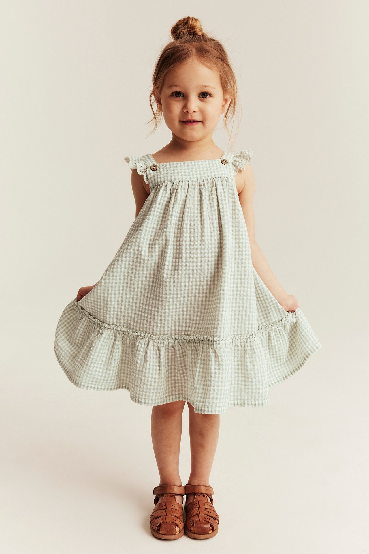 Front view: Kid wears a green checked woven dress with ruffled shoulders, a ruffled skirt, and brown sandals.