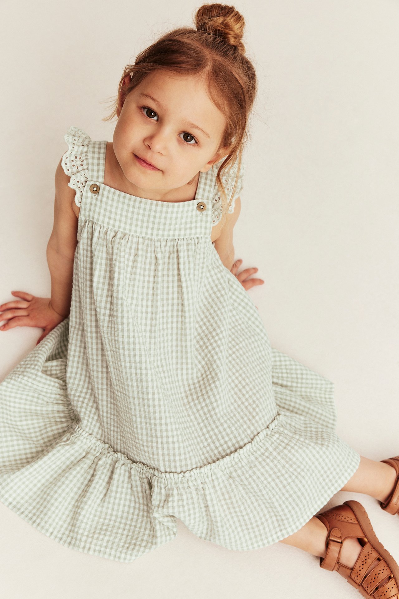 High-angle front view of a kid in a light green gingham cotton dress with ruffled shoulders and brown sandals.
