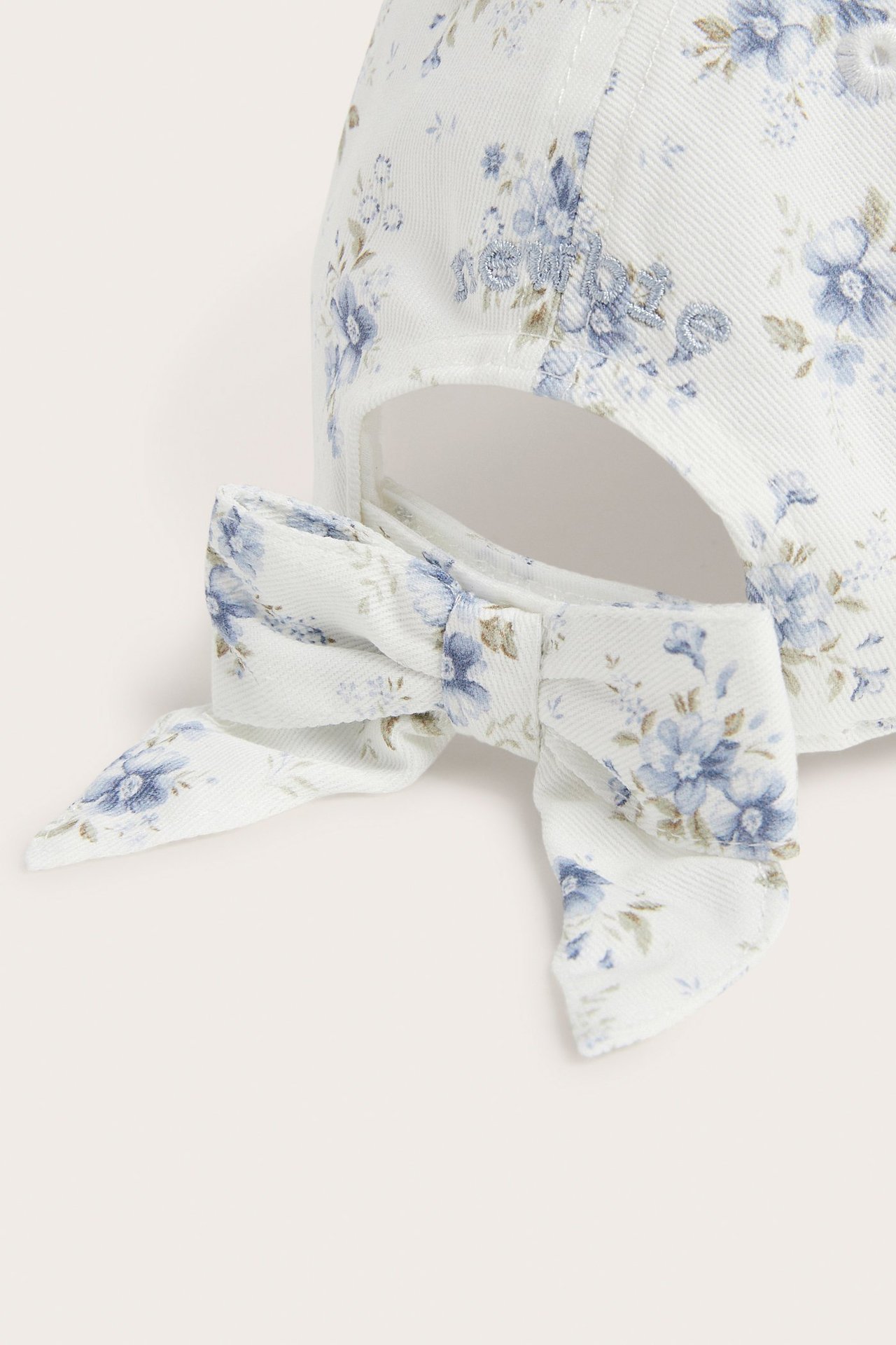 Close-up back view of an off-white cap with a blue floral pattern and a decorative bow.
