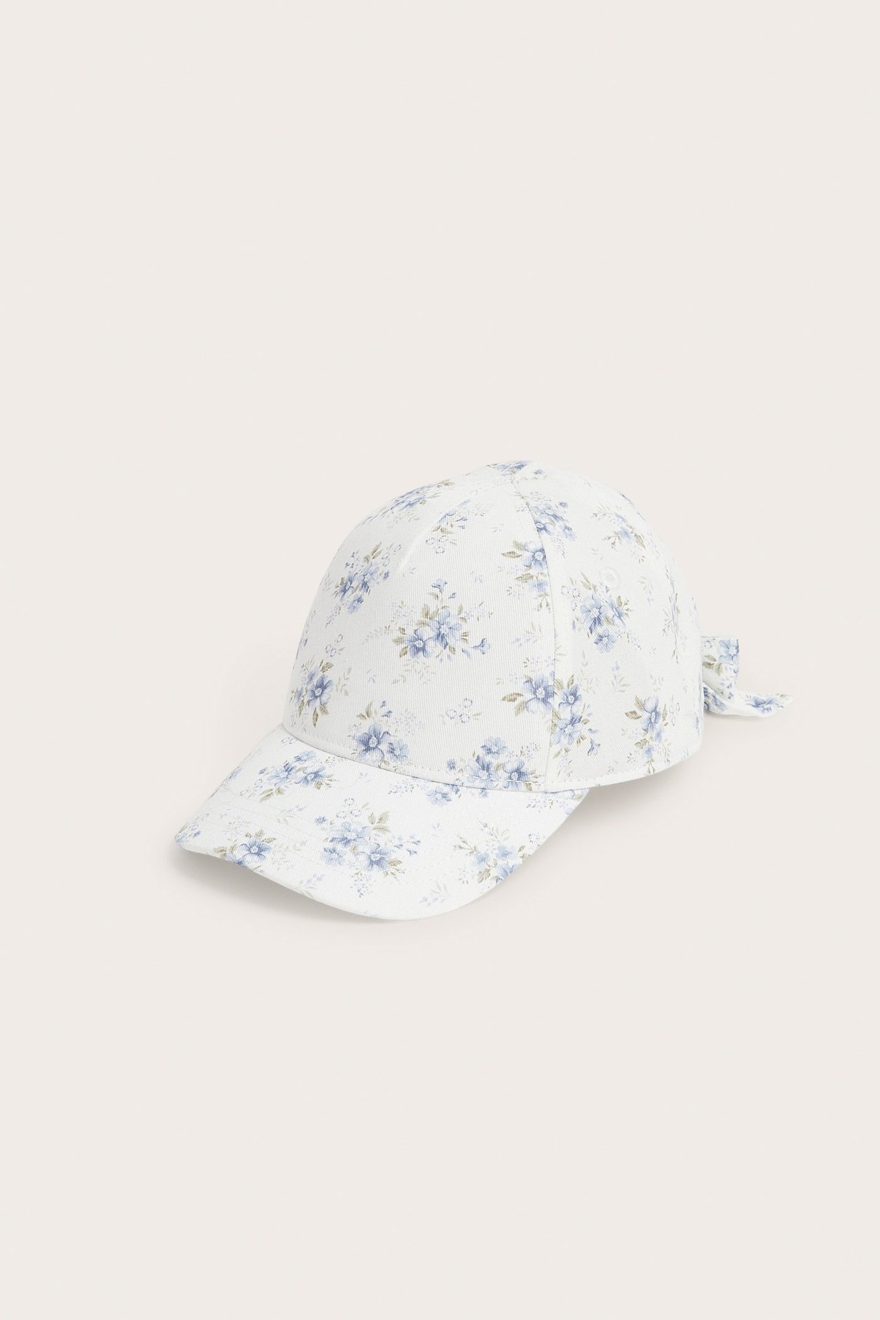 Side-front view of an off-white kid's cap with blue floral pattern, an adjustable velcro strap, and a decorative bow.
