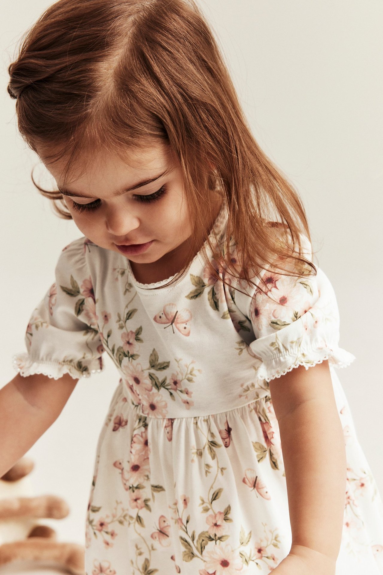 Nahaufnahme vorne eines Kindes in einem cremeweißen Baumwolljerseykleid mit Blumen- und Schmetterlingsmuster und Puffärmeln.