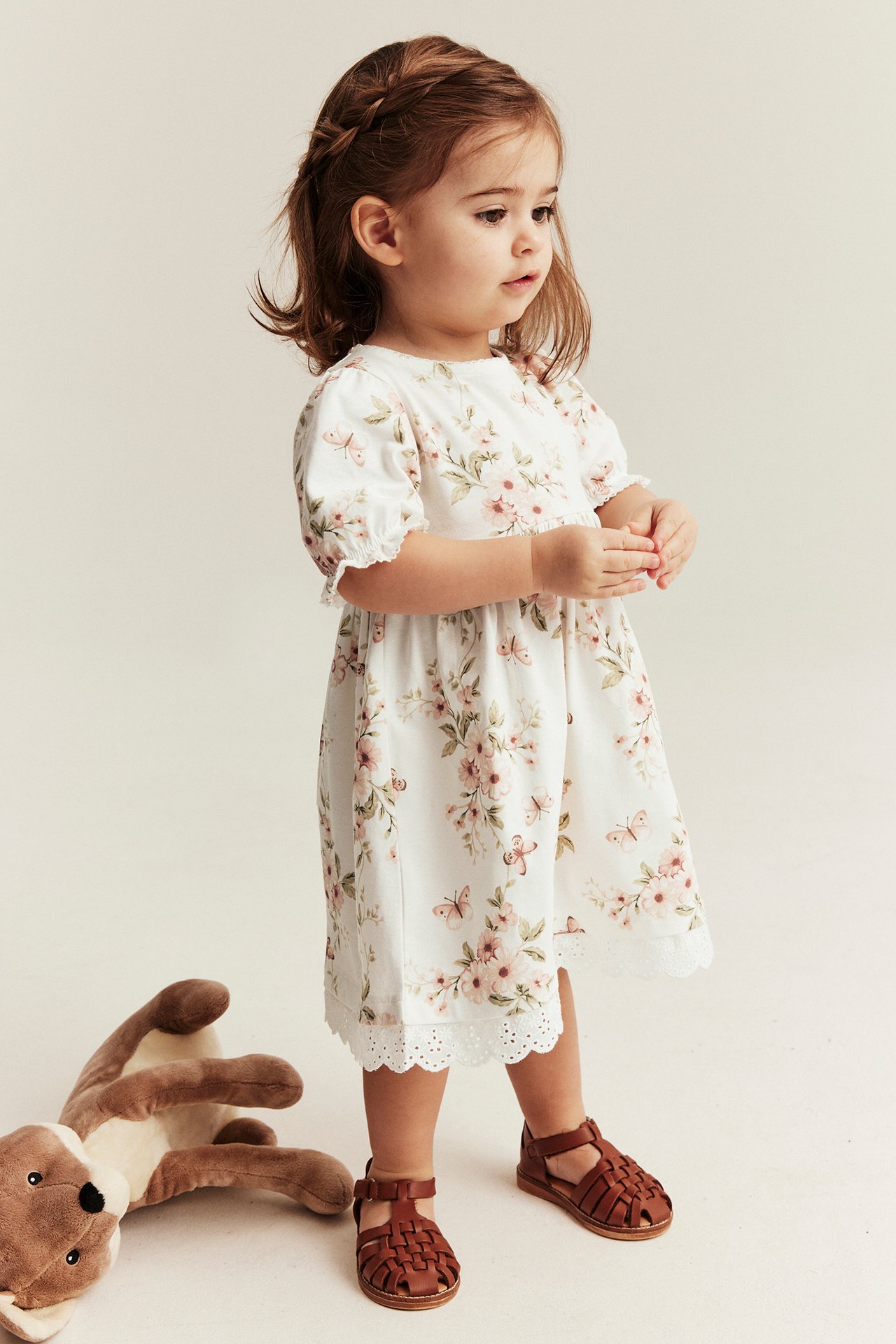 Front-side view: Kid in an offwhite cotton floral butterfly dress with puff sleeves and brown woven sandals.