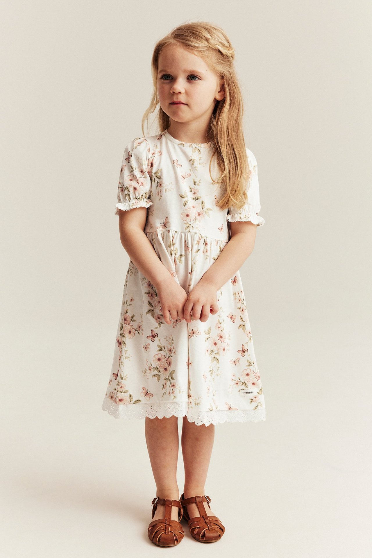 Front view of a kid wearing an off-white floral cotton jersey dress with puff sleeves, lace hem, and brown sandals.