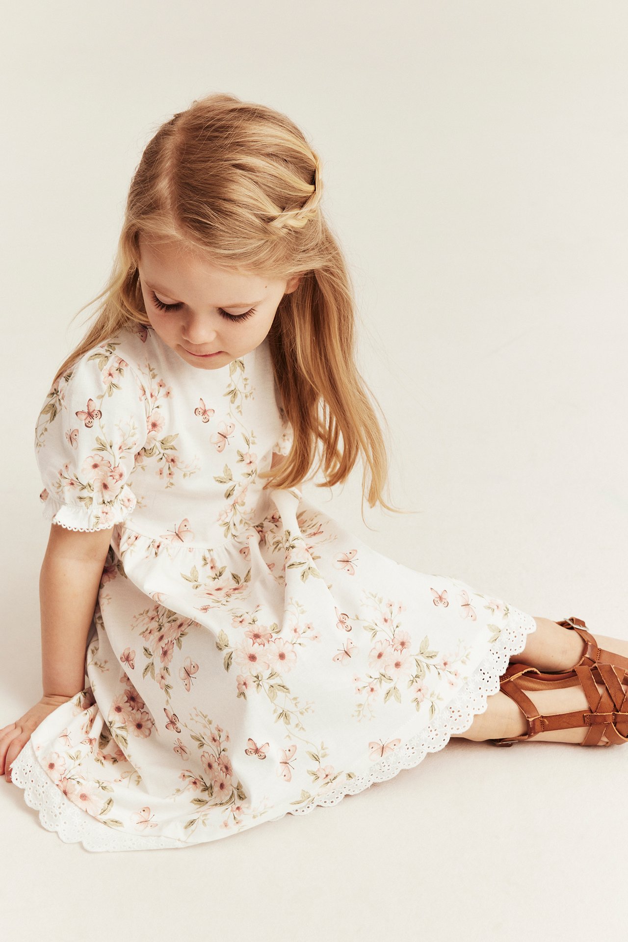 Mädchen trägt ein cremeweißes geblümtes Baumwollkleid mit Puffärmeln, Spitzenborte und braunen Sandalen, Vorderansicht.