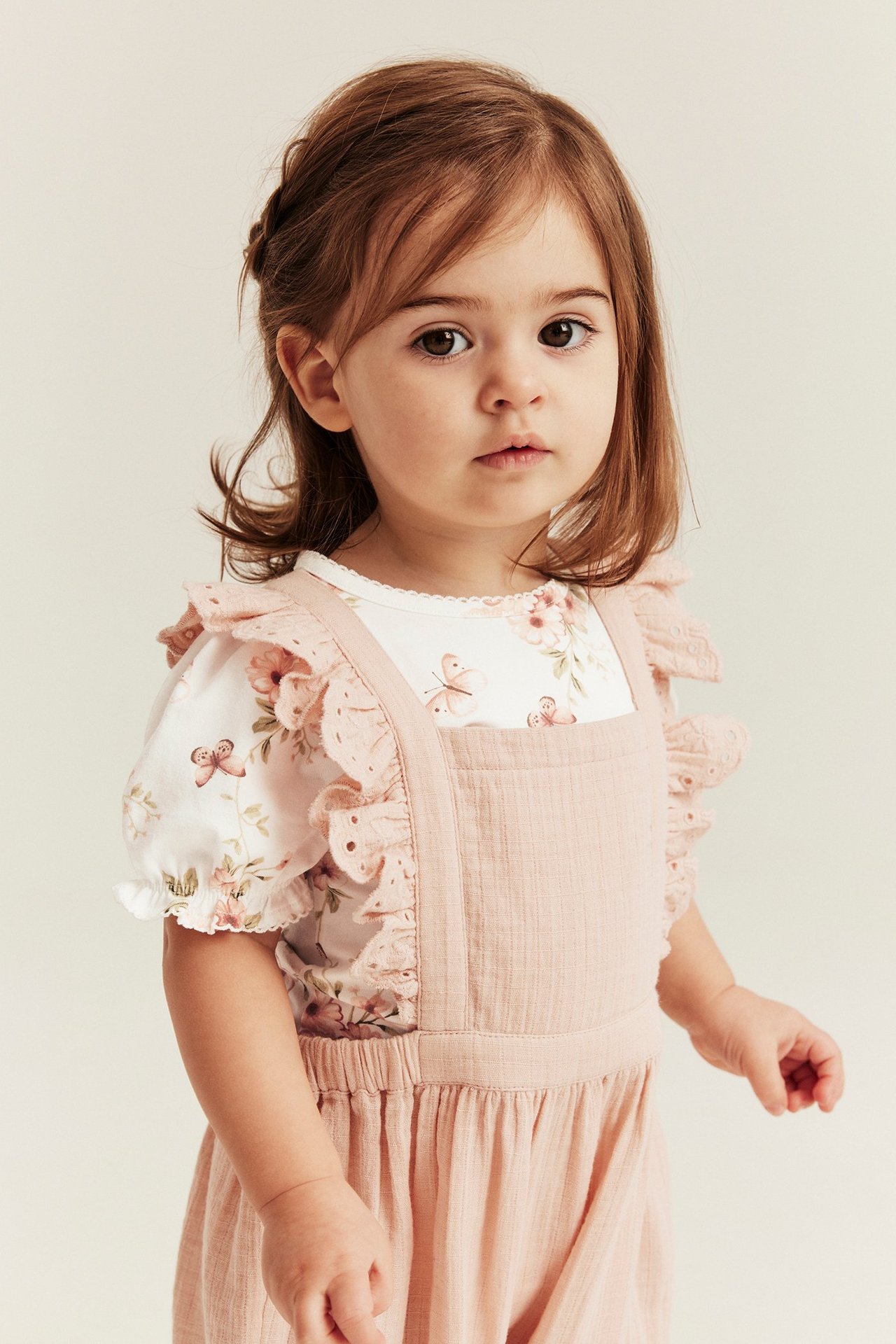 Vorderansicht: Baby in cremefarbenem Blumen-Body mit Puffärmeln unter rosa Rüschenoverall.