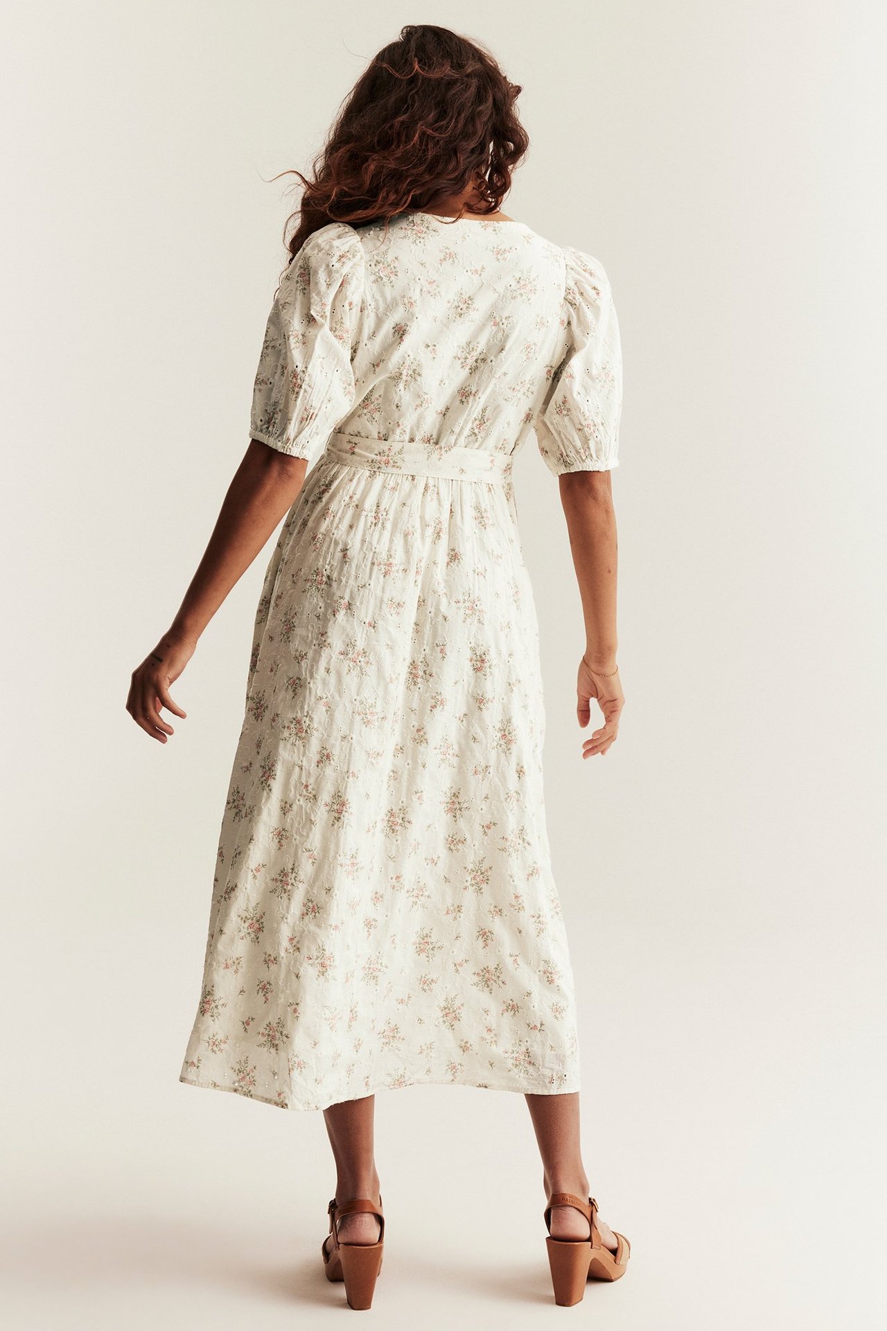 Rückenansicht einer Frau in off-white geblümtem Baumwollkleid mit Stickereien, Puffärmeln und braunen Keilsandalen.