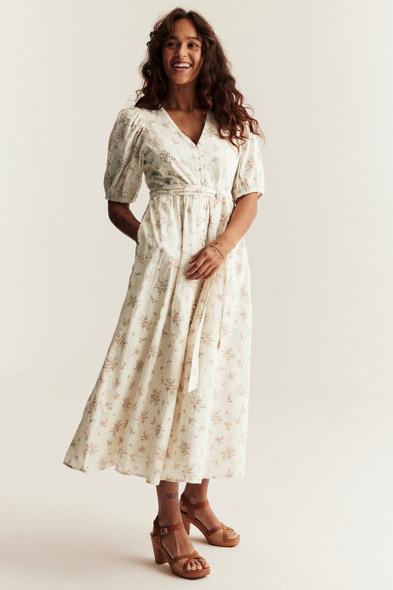 Frontansicht einer Frau in einem cremeweißen geblümten Baumwoll-Midikleid mit Puffärmeln und braunen Blockabsatz-Sandalen.