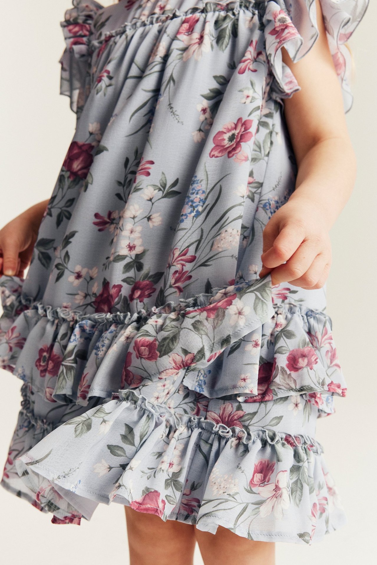 Close-up of a kid wearing a light blue floral chiffon dress with ruffled sleeves and tiered skirt.