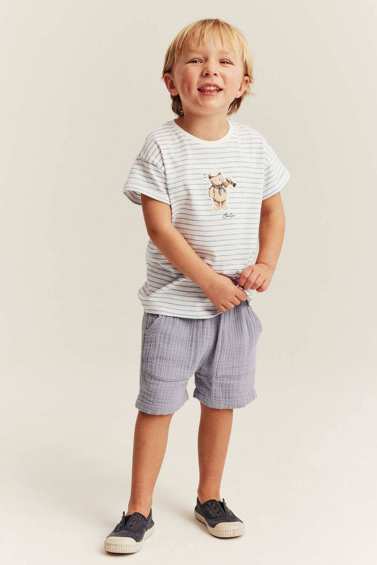 Vorderansicht eines Kindes in off-white gestreiftem Jersey-T-Shirt mit Teddy, hellblauen Shorts und dunklen Schuhen.