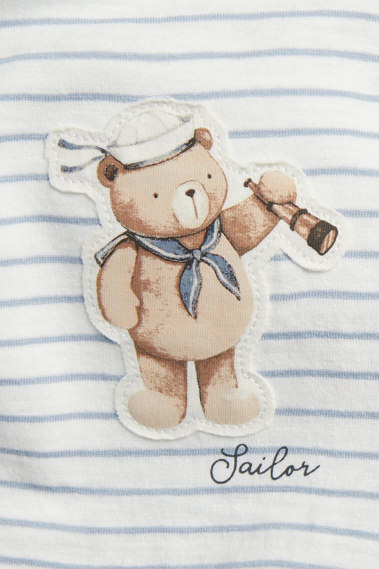 Close-up of an off-white striped cotton t-shirt with a sailor teddy bear application and "Sailor" text.
