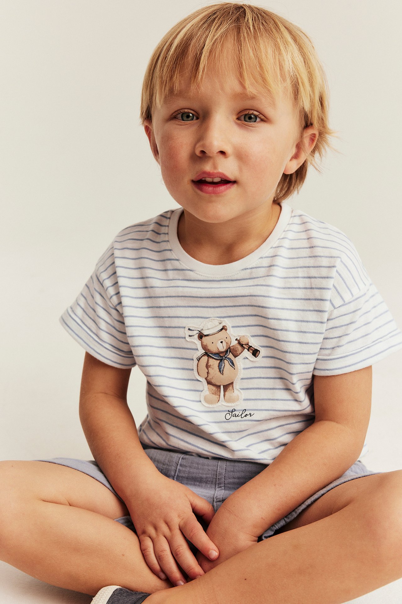 Front view of a kid in an off-white striped t-shirt with a sailor bear application and blue shorts.