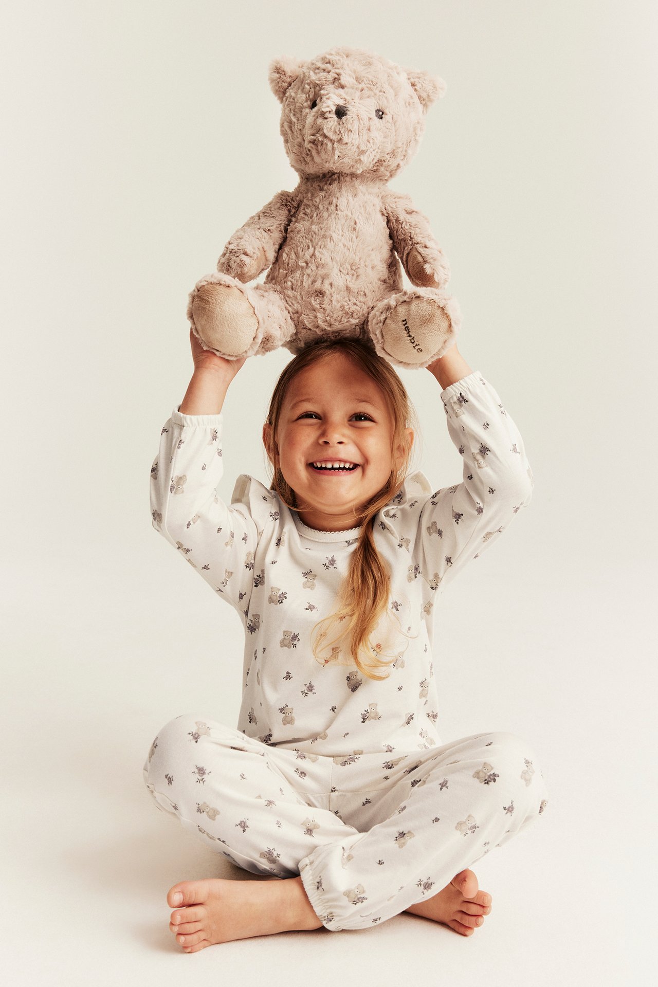 Vorderansicht eines Kindes in cremeweiß gemustertem Pyjama-Set, das einen beigen flauschigen Teddybär hält.