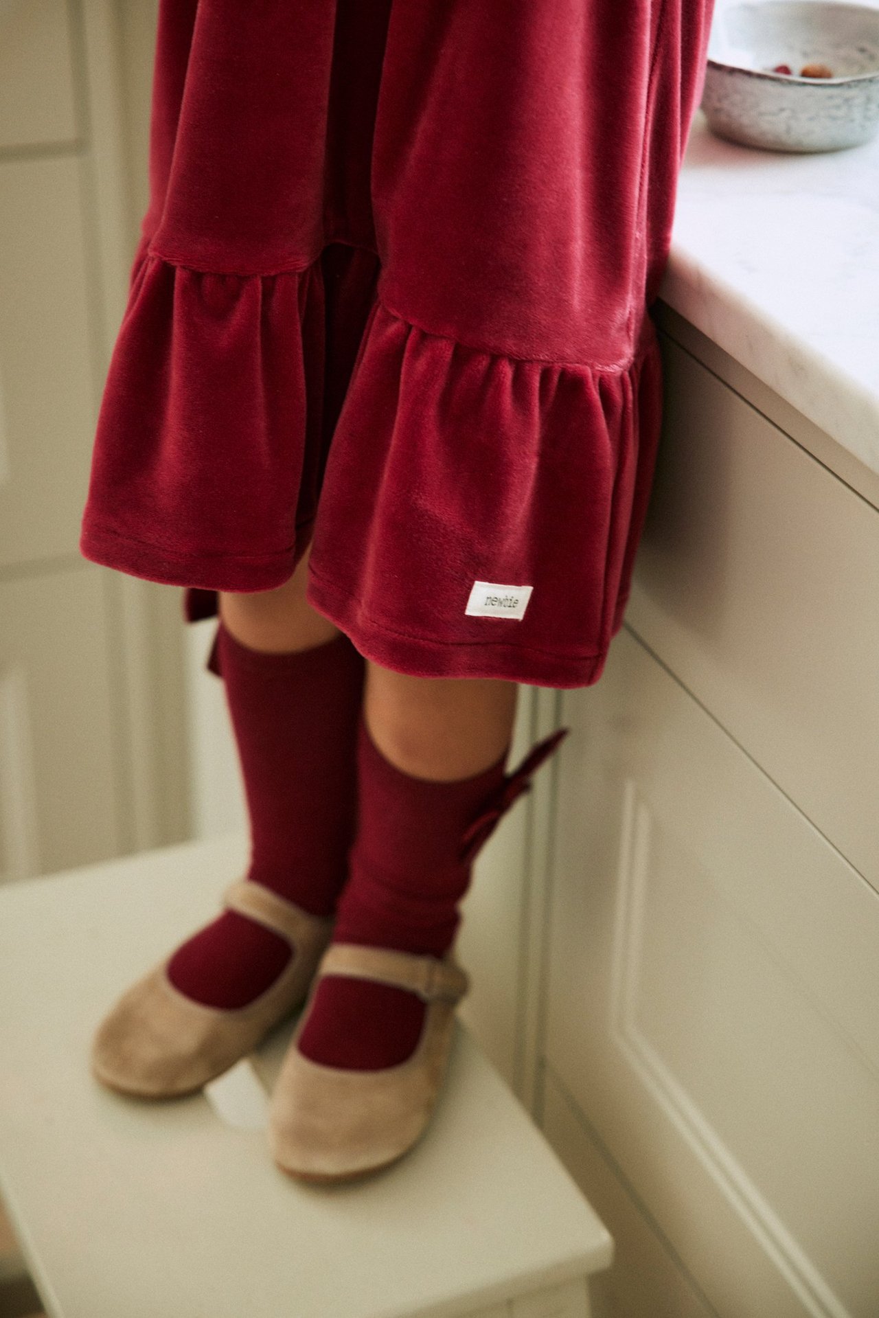 Vorderansicht: Kind trägt rote Kniestrümpfe mit Satinschleifen, rotes Samtkleid, beige Mary Jane Schuhe.