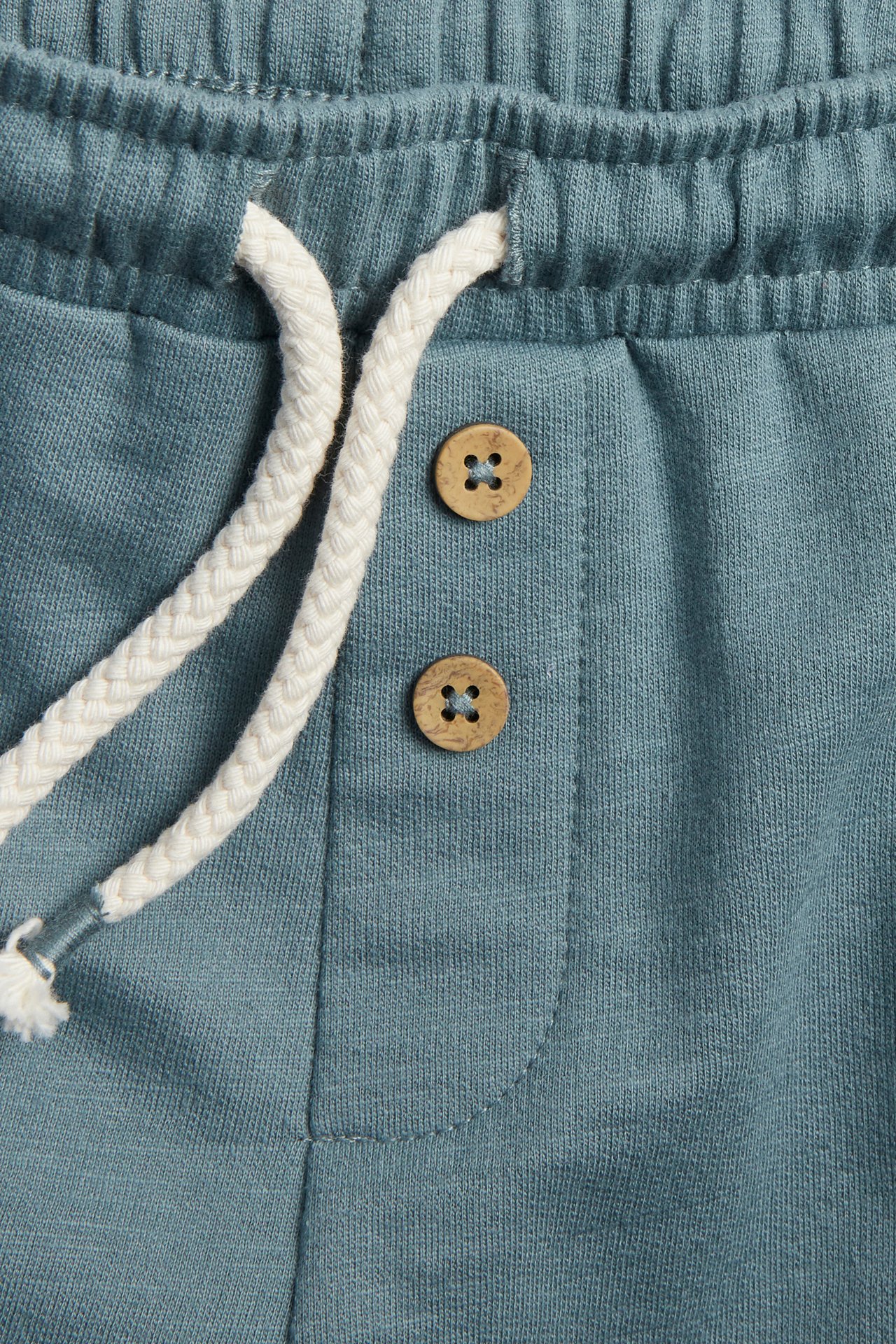 Close-up of dark turquoise slub jersey shorts with elastic waist, white drawstring, and two wooden buttons.