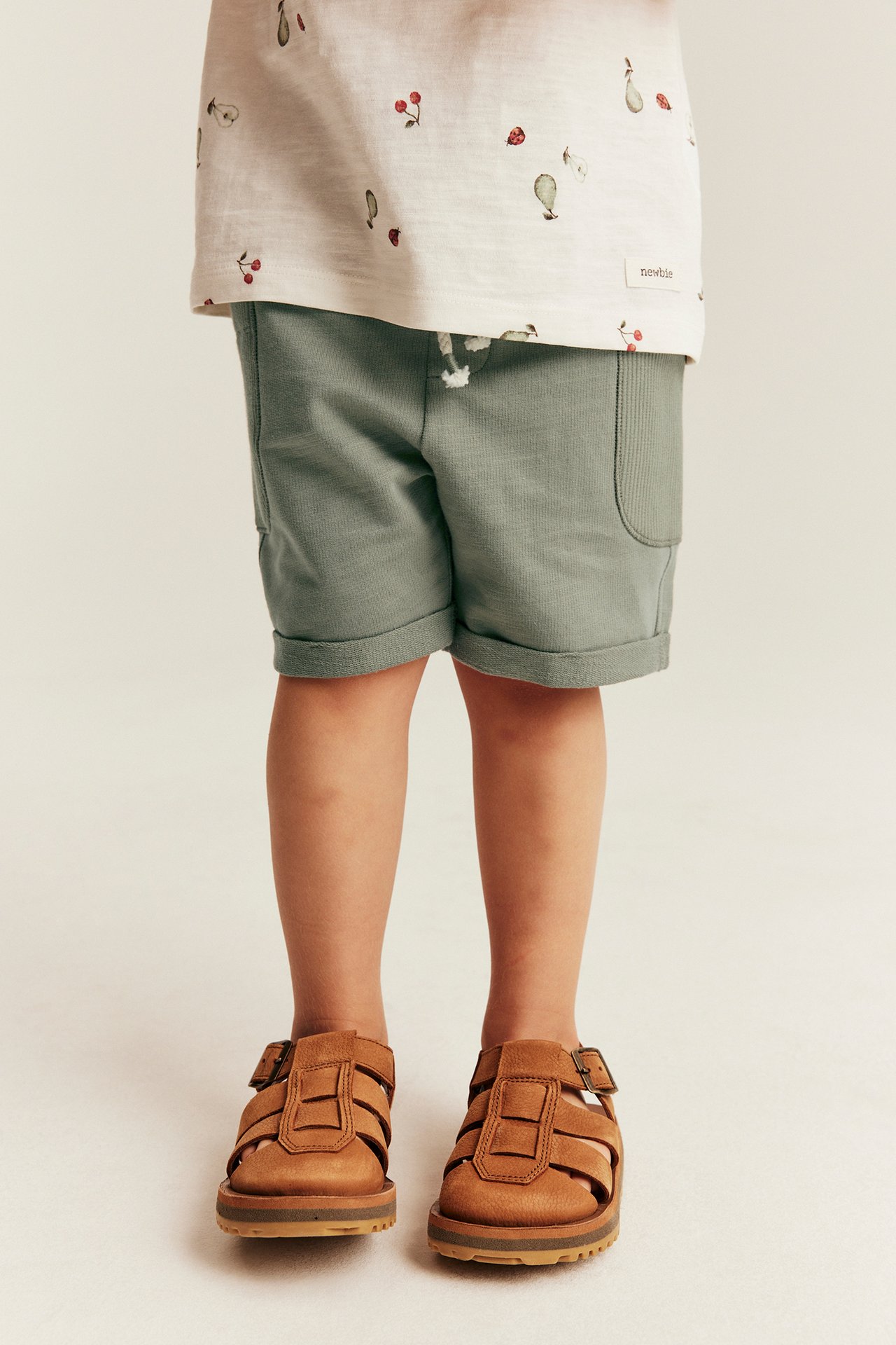 Front view of a kid wearing dark green slub jersey shorts with rolled-up cuffs, a patterned t-shirt, and brown sandals.