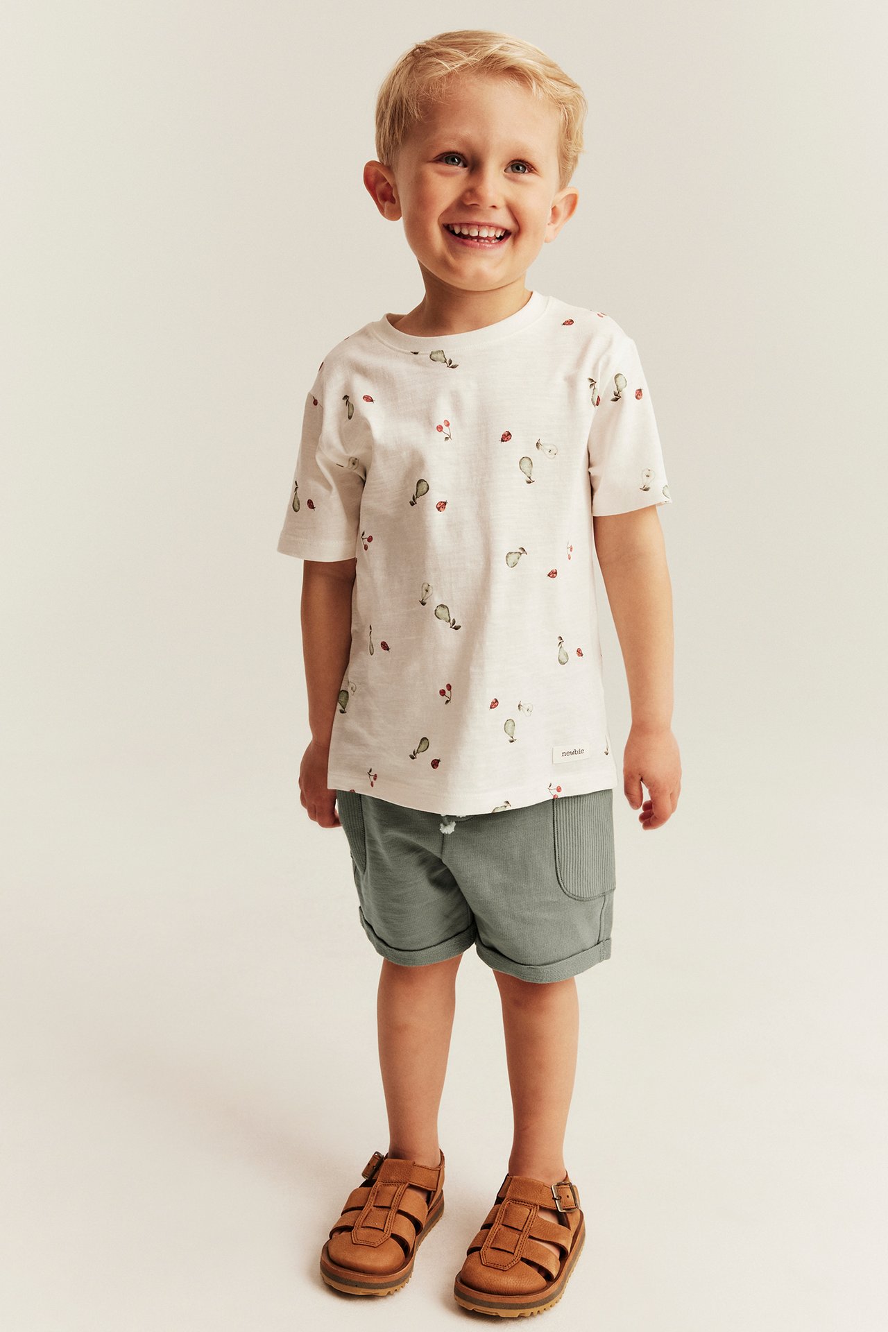 Front view: Kid wears dark green slub jersey shorts, a white patterned t-shirt, and brown sandals.