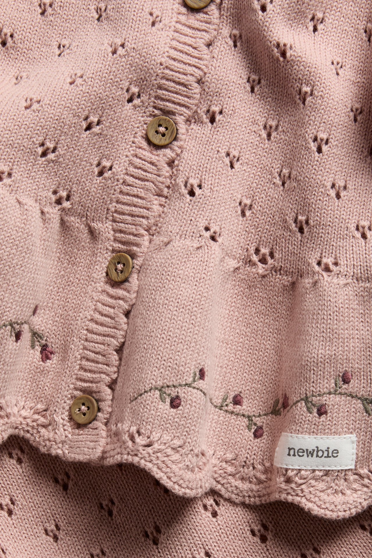 Close-up of a pink knitted baby cardigan with eyelet pattern, scalloped edges, and berry embroidery.