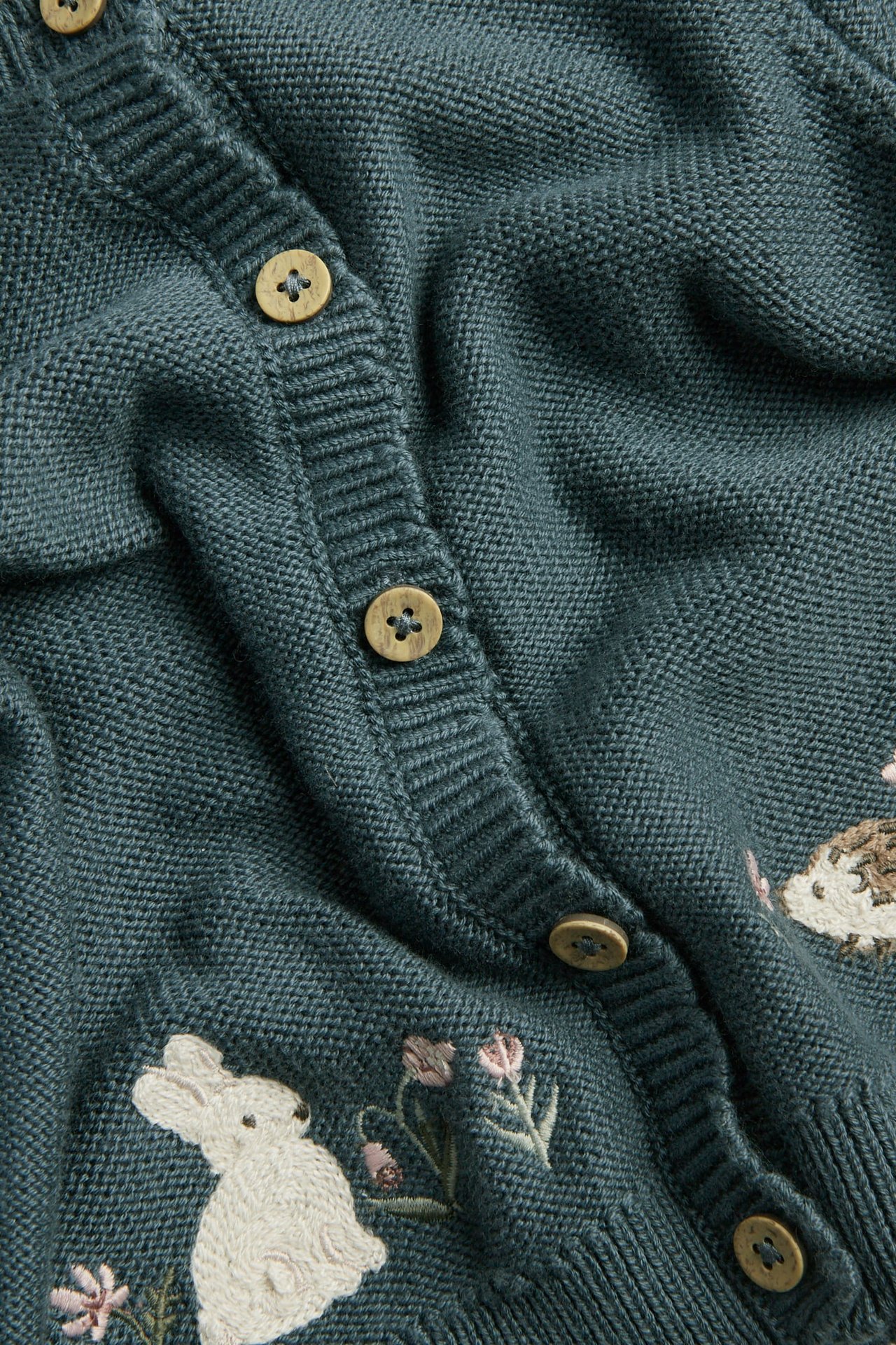 Nahaufnahme einer dunkelgrünen Strickjacke mit Holzknöpfen und besticktem Kaninchen, Igel und Blumen.