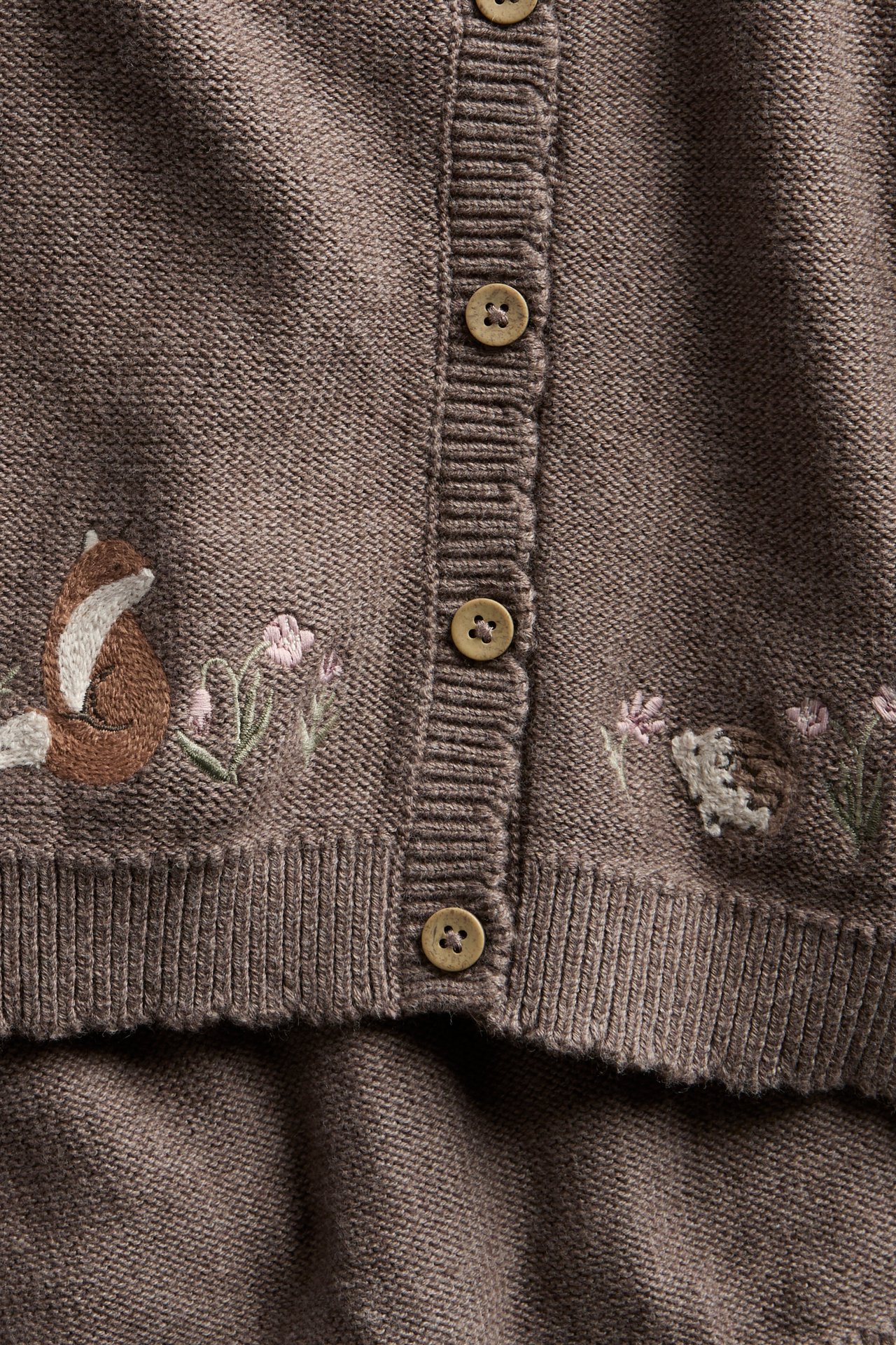 Close-up: brown knitted cardigan with embroidered fox, hedgehog, flowers, buttons, and matching knitted bottom.