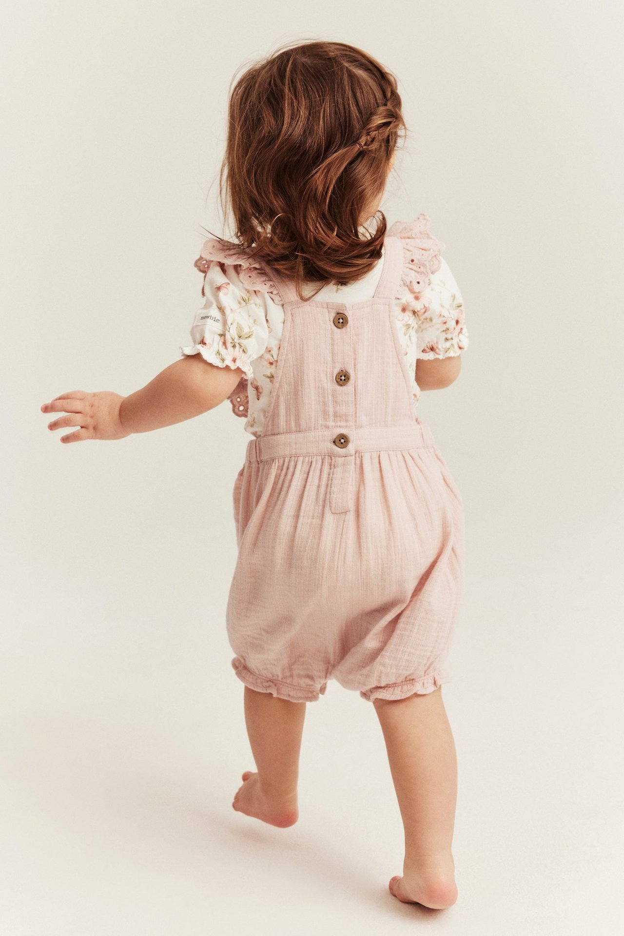 Rückenansicht eines Babys in hellrosa Baumwoll-Latzshorts mit Spitzenrüschen über einem geblümten Oberteil.