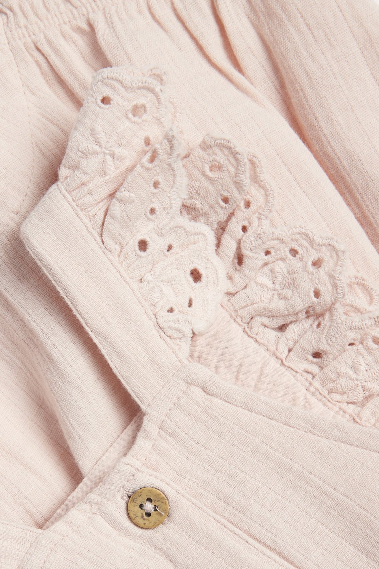 Nahaufnahme vorne: Hellrosa gewebte Baumwoll-Latzshorts mit Spitzenrüschen an den Trägern und einem Holzknopf.