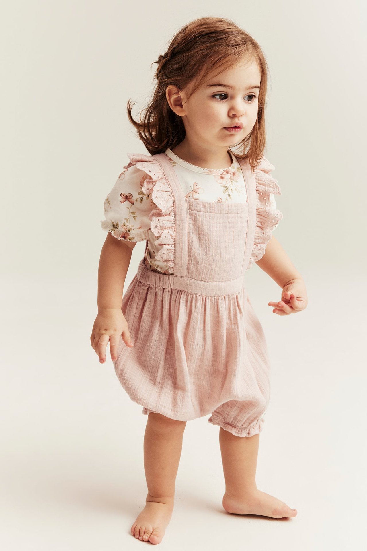 Front view of a baby girl wearing light pink woven overall shorts with ruffled straps over a white floral top.