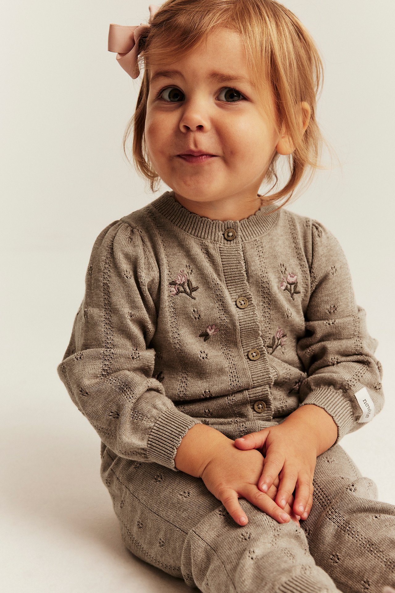Front view of a baby girl wearing a light brown knitted cardigan with pink floral embroidery and matching knitted pants.