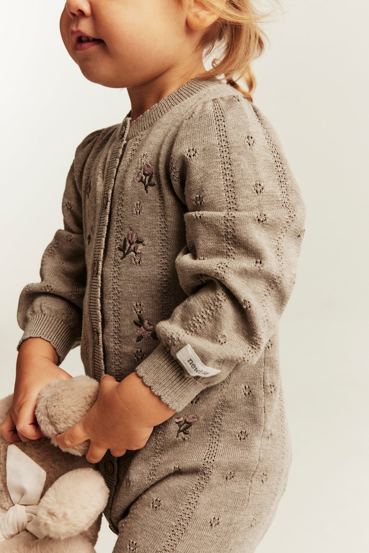 Side view of a kid in a taupe knitted floral romper with eyelet details, holding a plush toy.