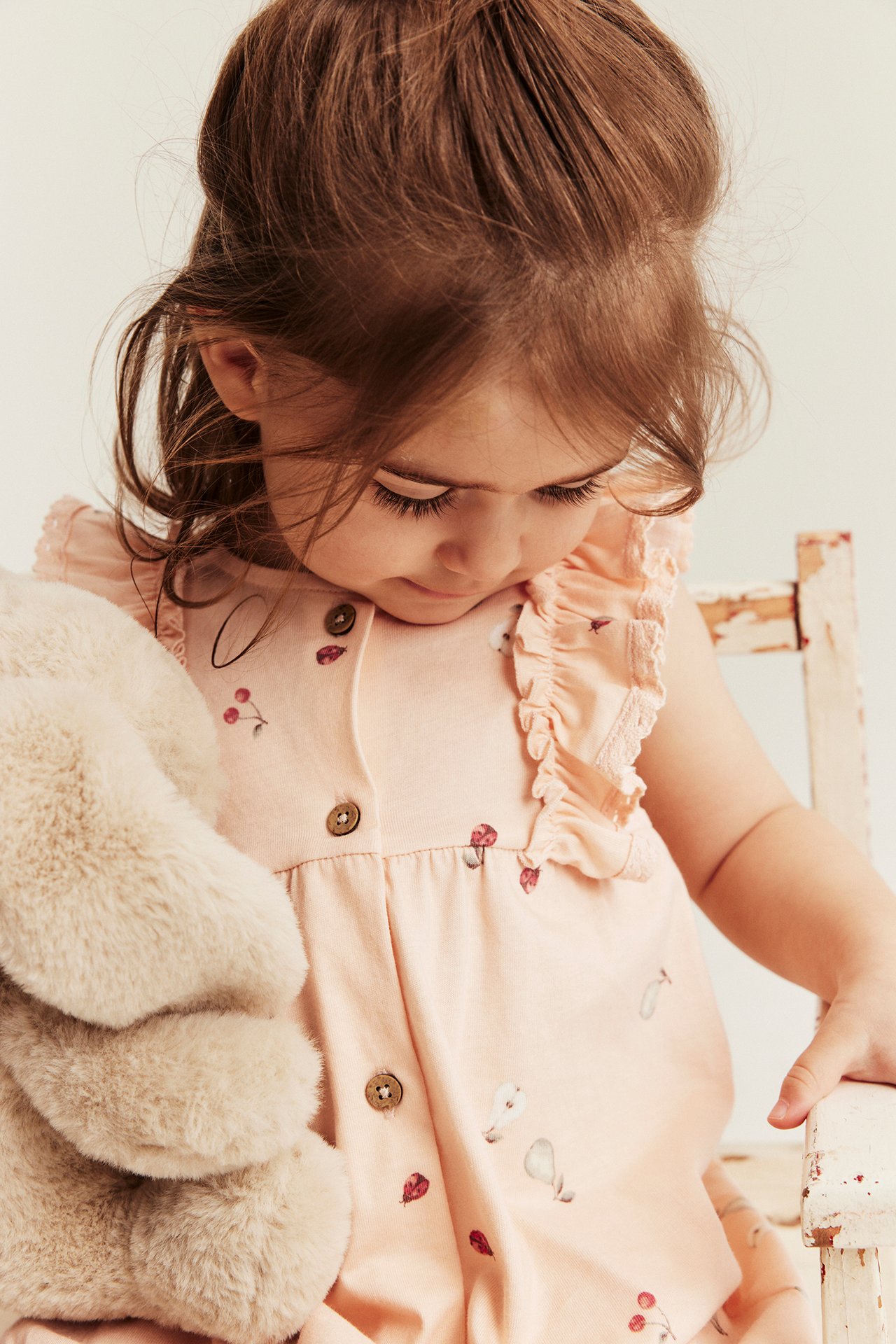Close-up of a kid wearing a light pink patterned cotton romper with ruffles, holding a beige fluffy toy.