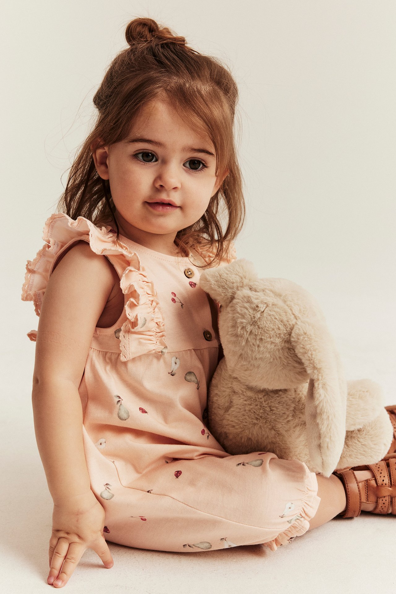 Front view of a baby in a light pink fruit-print sleeveless romper, holding a fluffy beige bunny. Brown sandals.