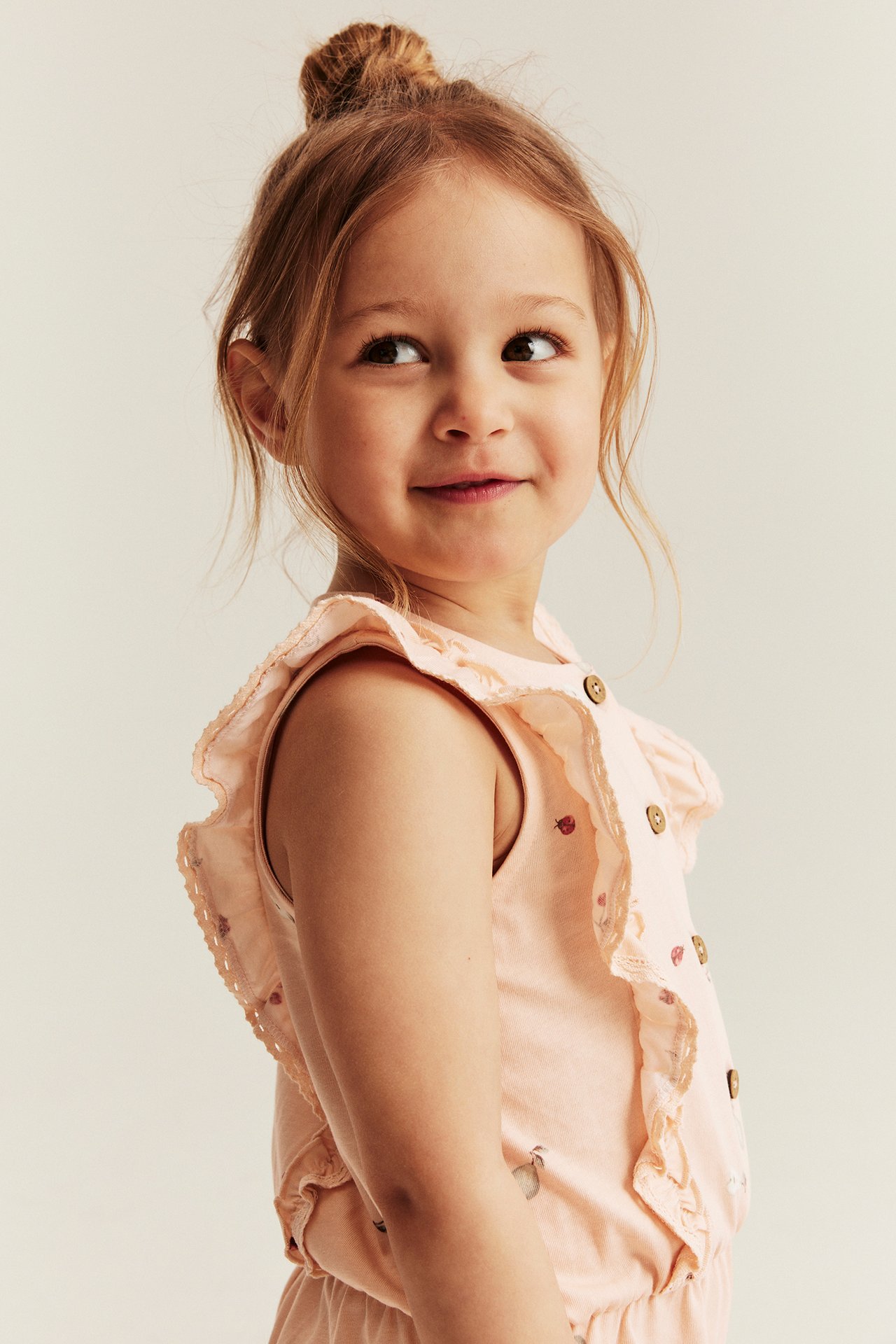 Close-up: Kid in light orange patterned cotton jumpsuit with ruffles, lace, and front buttons.