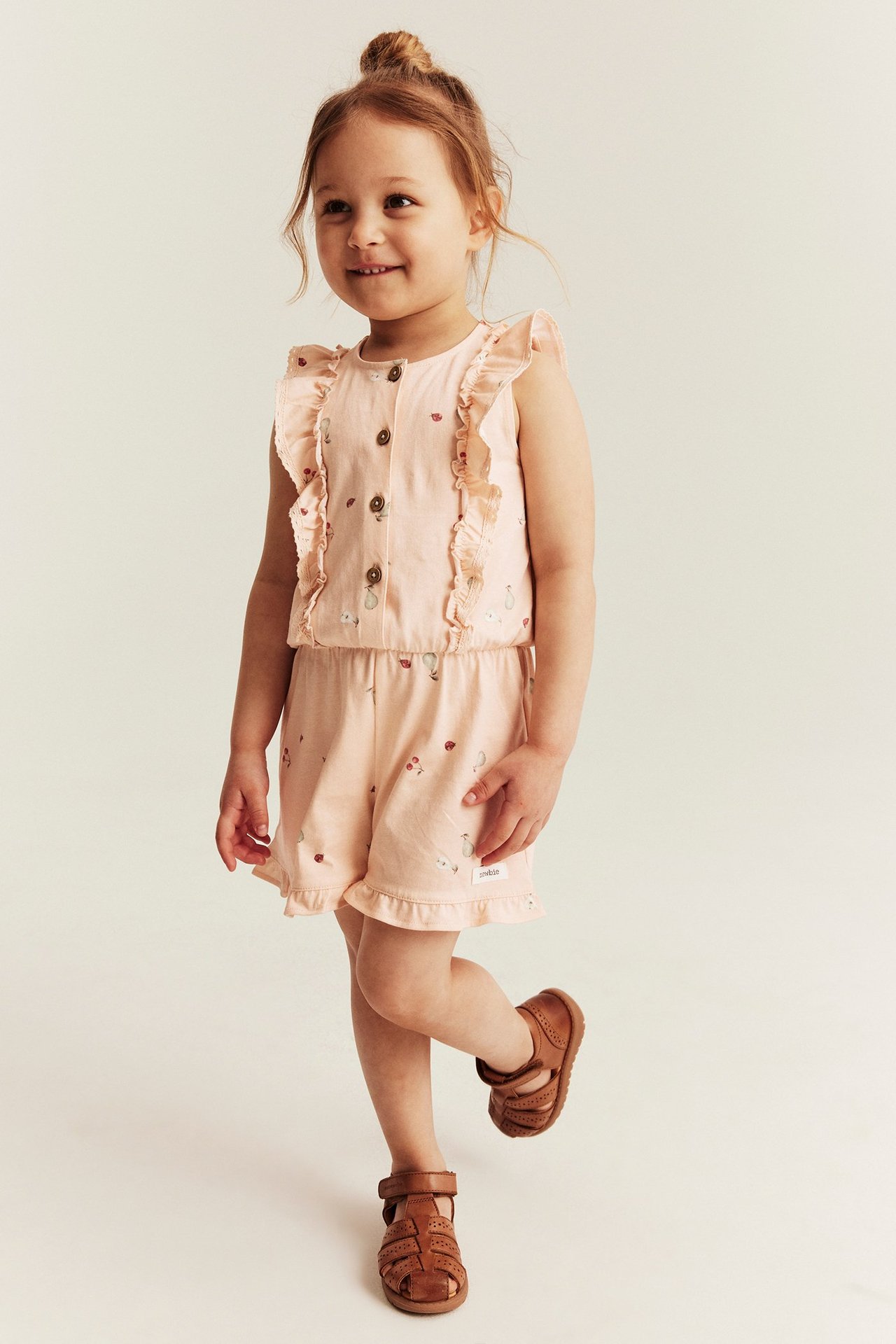 Vorderansicht: Kind in hell-pfirsichfarbenem gemustertem Jersey-Jumpsuit mit Rüschen und Knöpfen, braune Sandalen tragend.