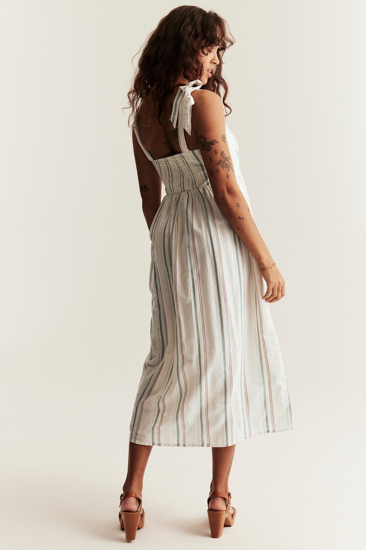 Side view of a woman in an off-white striped cotton midi dress with tie shoulder straps and brown sandals.