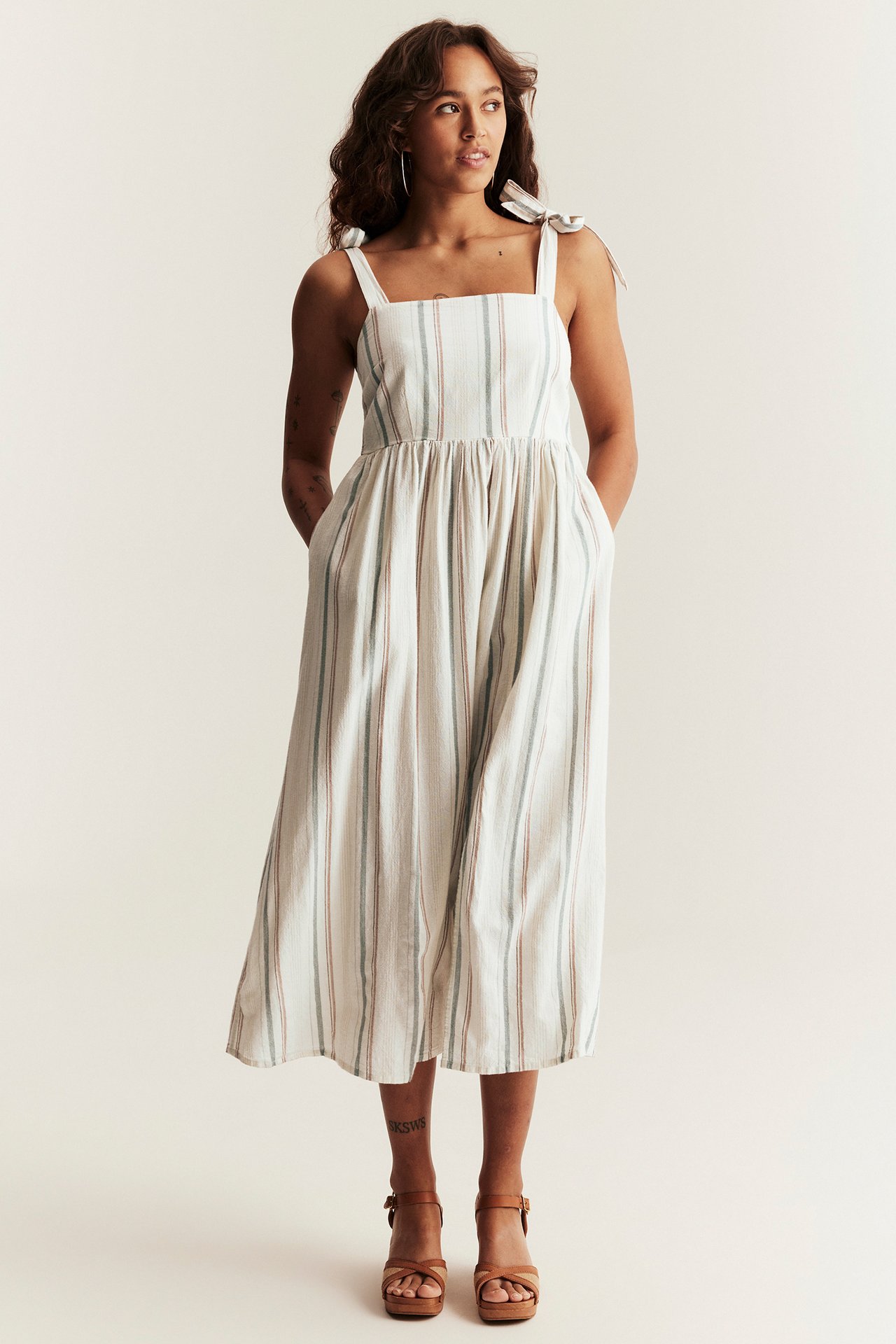 Vorderansicht einer Frau in einem gestreiften, cremefarbenen Baumwollkleid mit Bindeträgern, Smok am Rücken und braunen Sandalen.