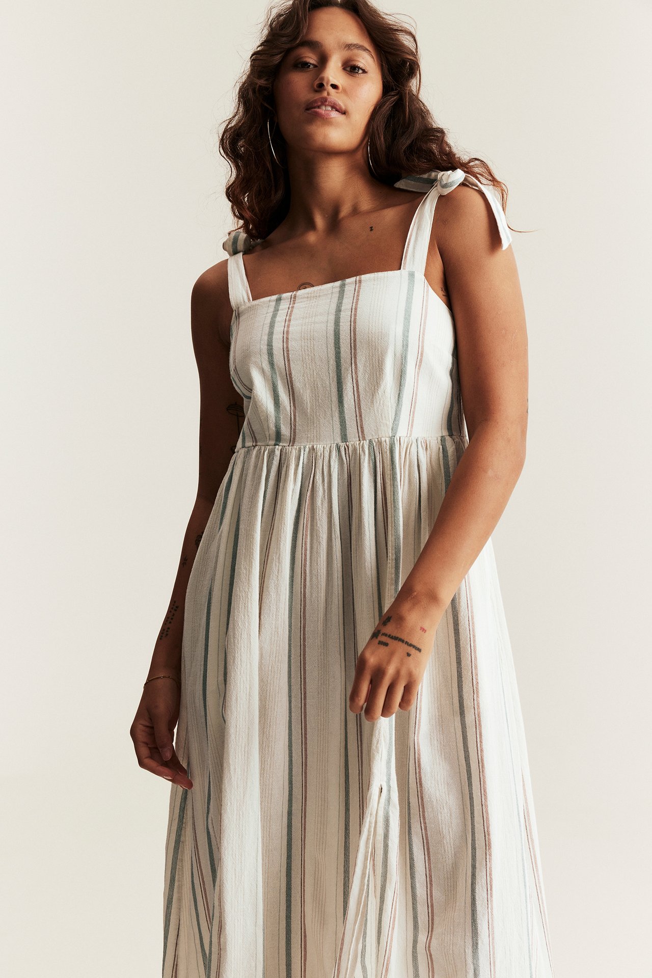 Front view of a woman wearing an offwhite striped woven cotton dress with tie-shoulder straps and a smocked back.