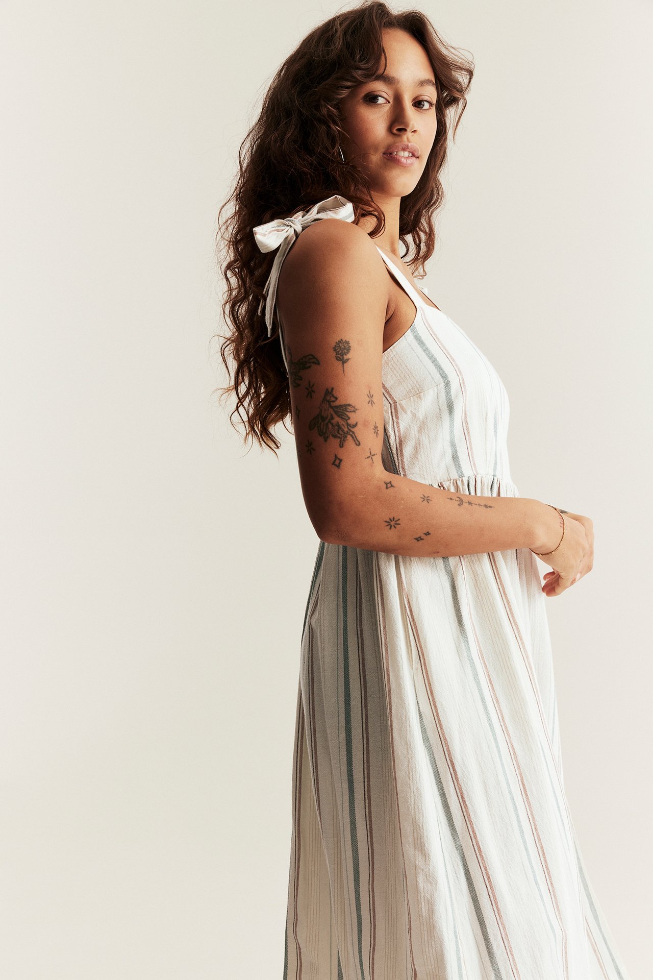 Seitenansicht einer Frau in einem cremeweißen gestreiften Baumwollkleid mit Schleifenträgern und Raffung an der Taille.