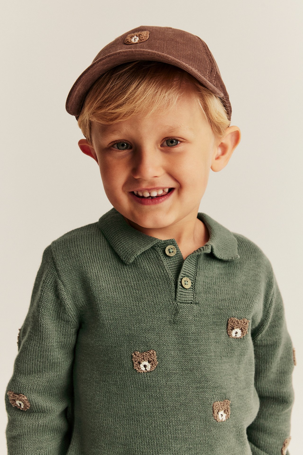 Vorderansicht eines Kindes in grünem Strick-Poloshirt mit Bärenstickerei und brauner Kordkappe.