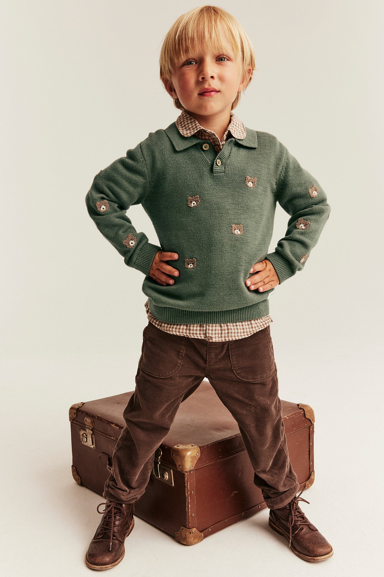 Front view: kid's green bear-embroidered sweater, plaid shirt, brown corduroy pants, and brown boots.